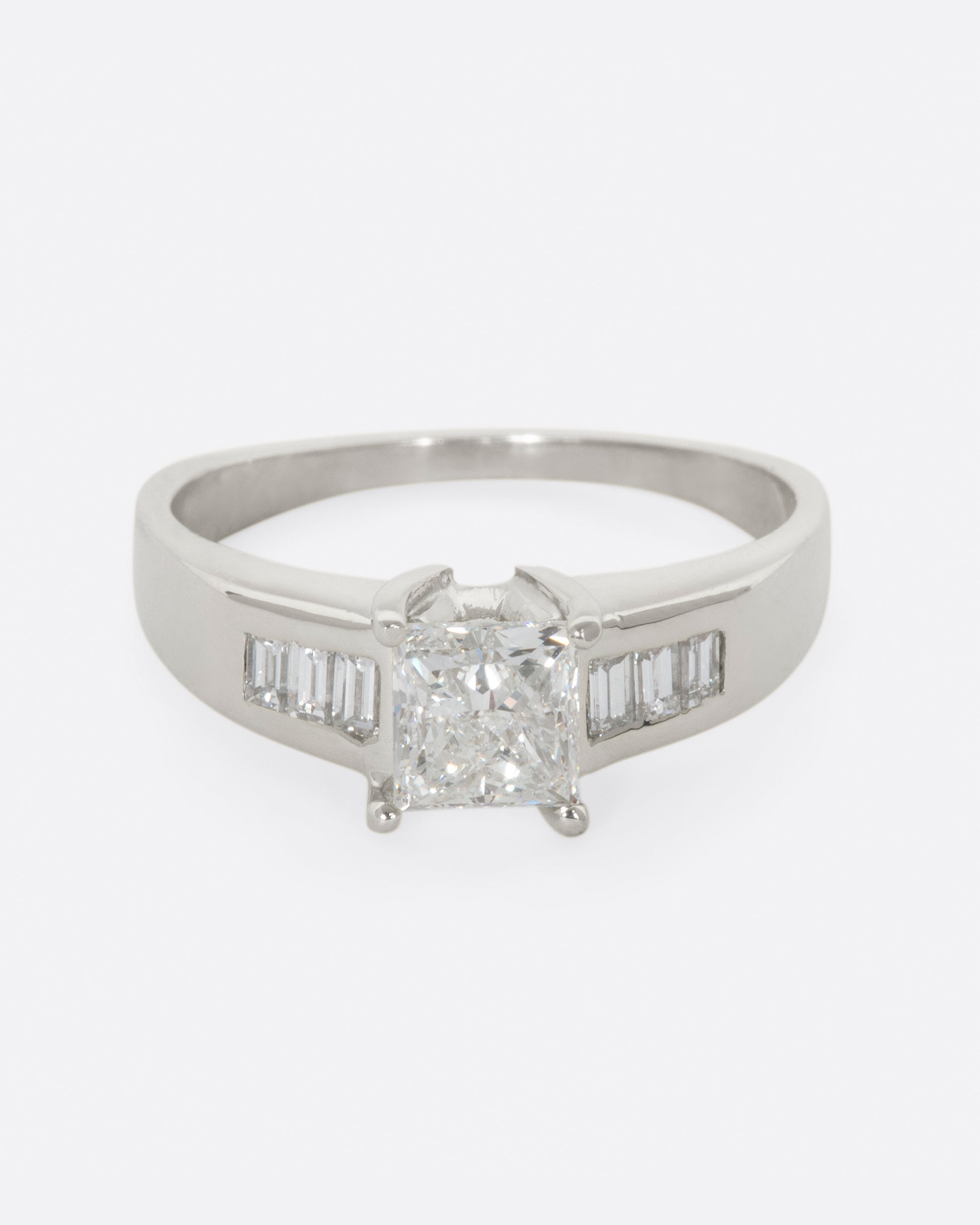 A platinum ring with a prong set princess cut center diamond and three baguette diamonds on either side. View from the front.