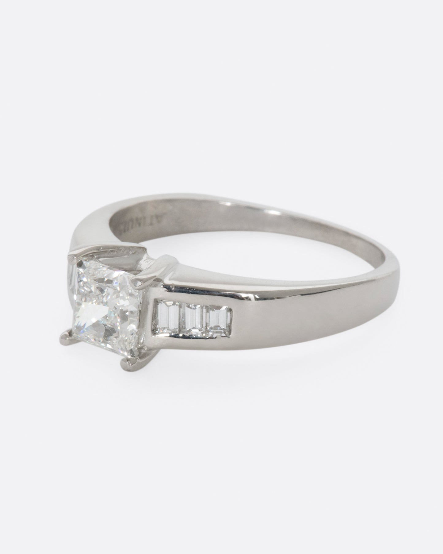 A platinum ring with a prong set princess cut center diamond and three baguette diamonds on either side. View from the left side.