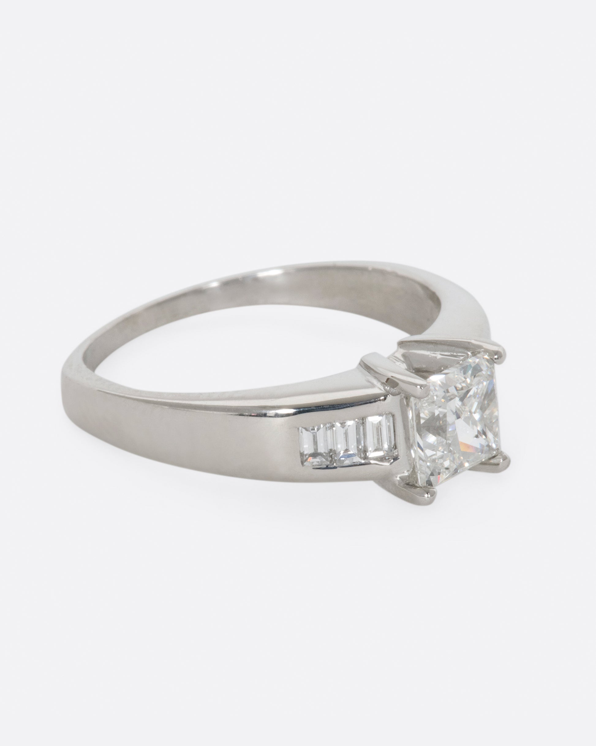 A platinum ring with a prong set princess cut center diamond and three baguette diamonds on either side. View from the right side.