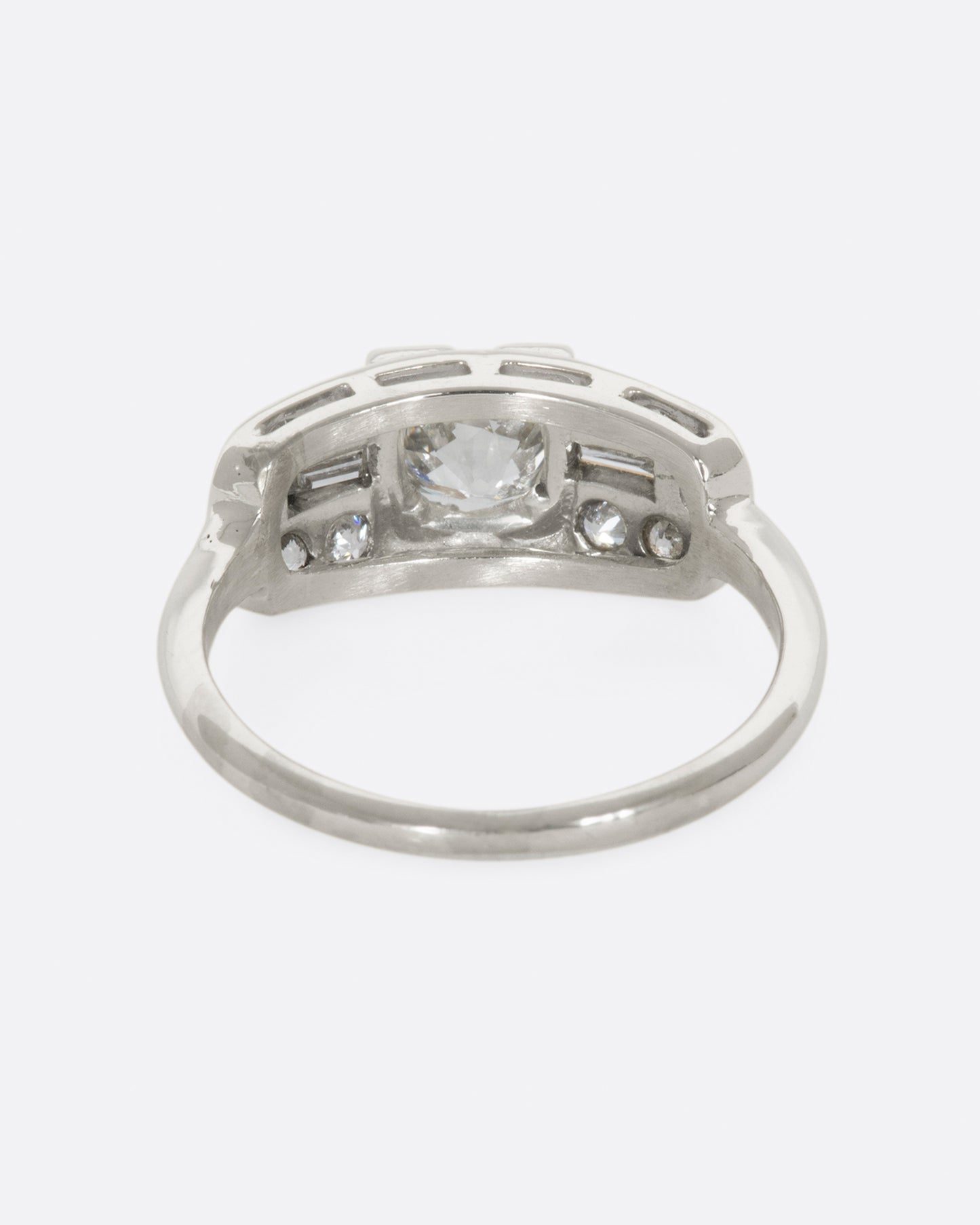 A white gold ring with round center diamond and baguette and round diamonds surrounding on a wide flat face. View from the back.