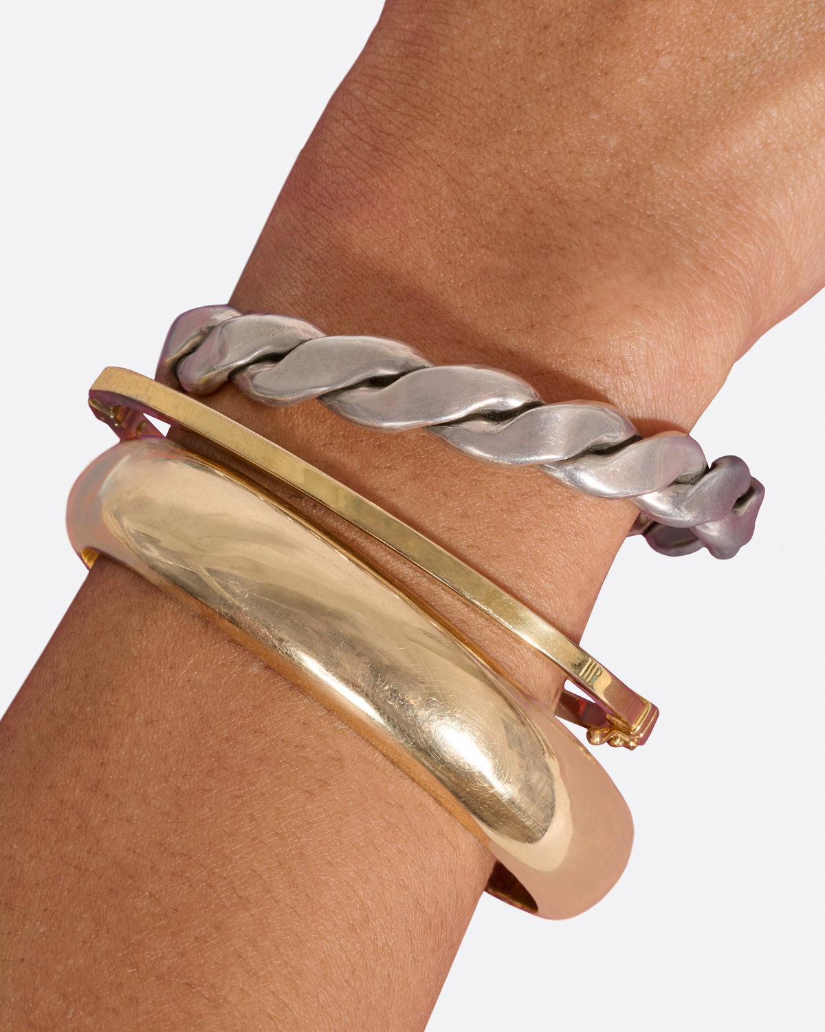A yellow gold oval hinged bangle with a security clasp, shown worn stacked with a twisted sterling cuff and a wider yellow gold bangle.