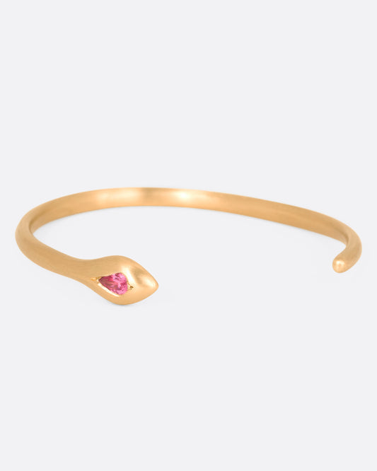 A matte yellow gold snake cuff bracelet with a pear shaped pink sapphire on its head, shown from the side.