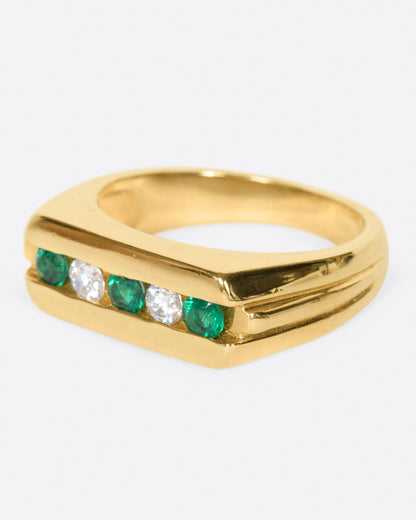A yellow gold ring with a row of five channel set stones; three emeralds and two diamonds. Shown from the side.
