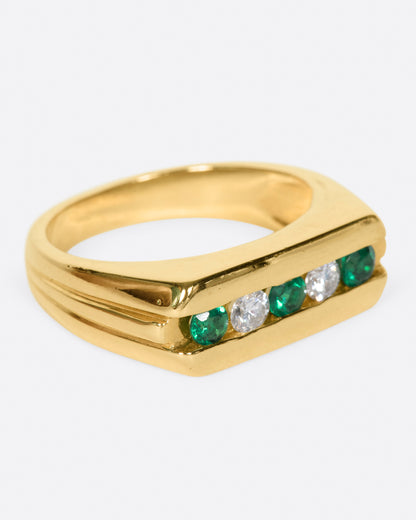 A yellow gold ring with a row of five channel set stones; three emeralds and two diamonds. Shown from the side.