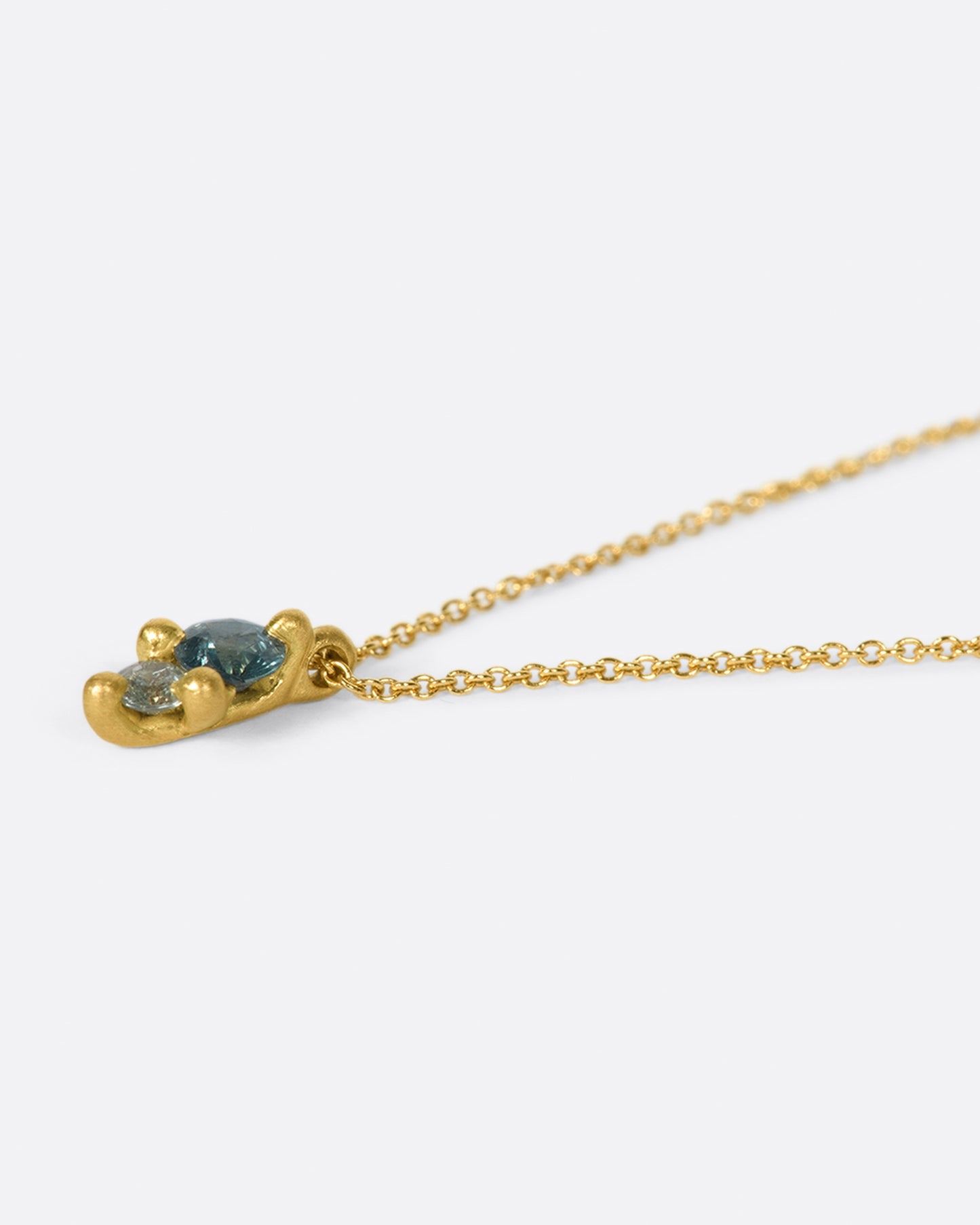 A pendant with two sapphires; one blue and one gray, set in matte gold prongs on a cable chain necklace.