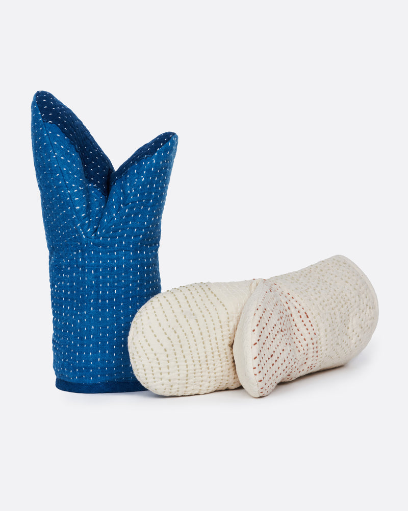 A pair of hand stitched oven mitts; one blue and one ivory. The blur is shown from the side standing up and the ivory is shown lying flat from the top.