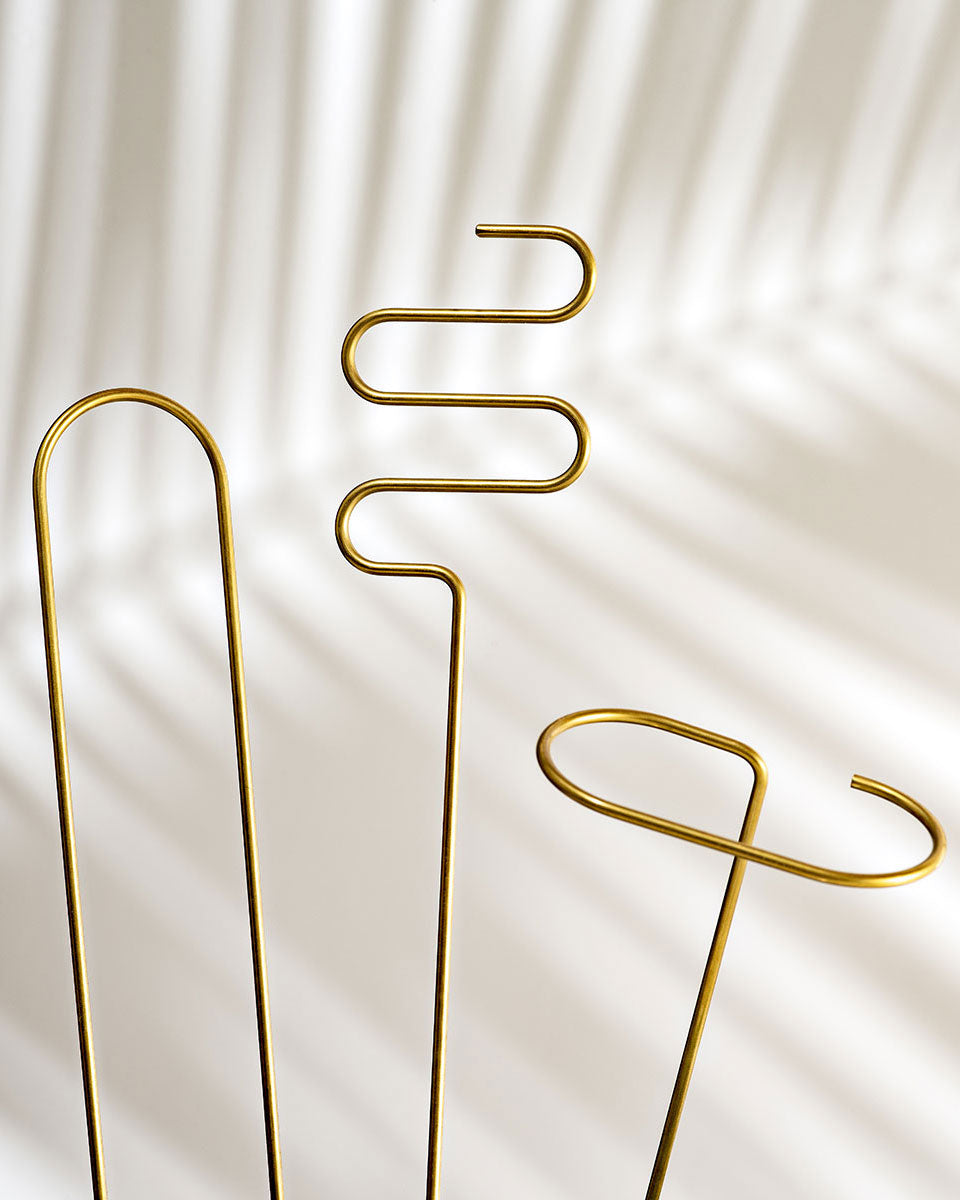 three brass plant sticks side by side on a white background, each one is bent into a different shape