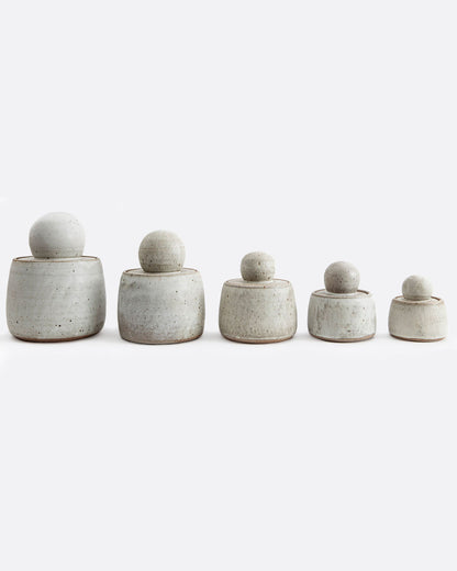 Five white stoneware jars with lids with ball shaped tops. Shown from biggest to smallest from the front.