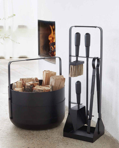 A black powder coated basket with leather on the handle and around the middle with pieces of wood inside. Shown from the front. Next to the basket is a black stand with four fireplace tools with black leather details on them. Shown from the front. There is a fire in a fireplace in the background. 