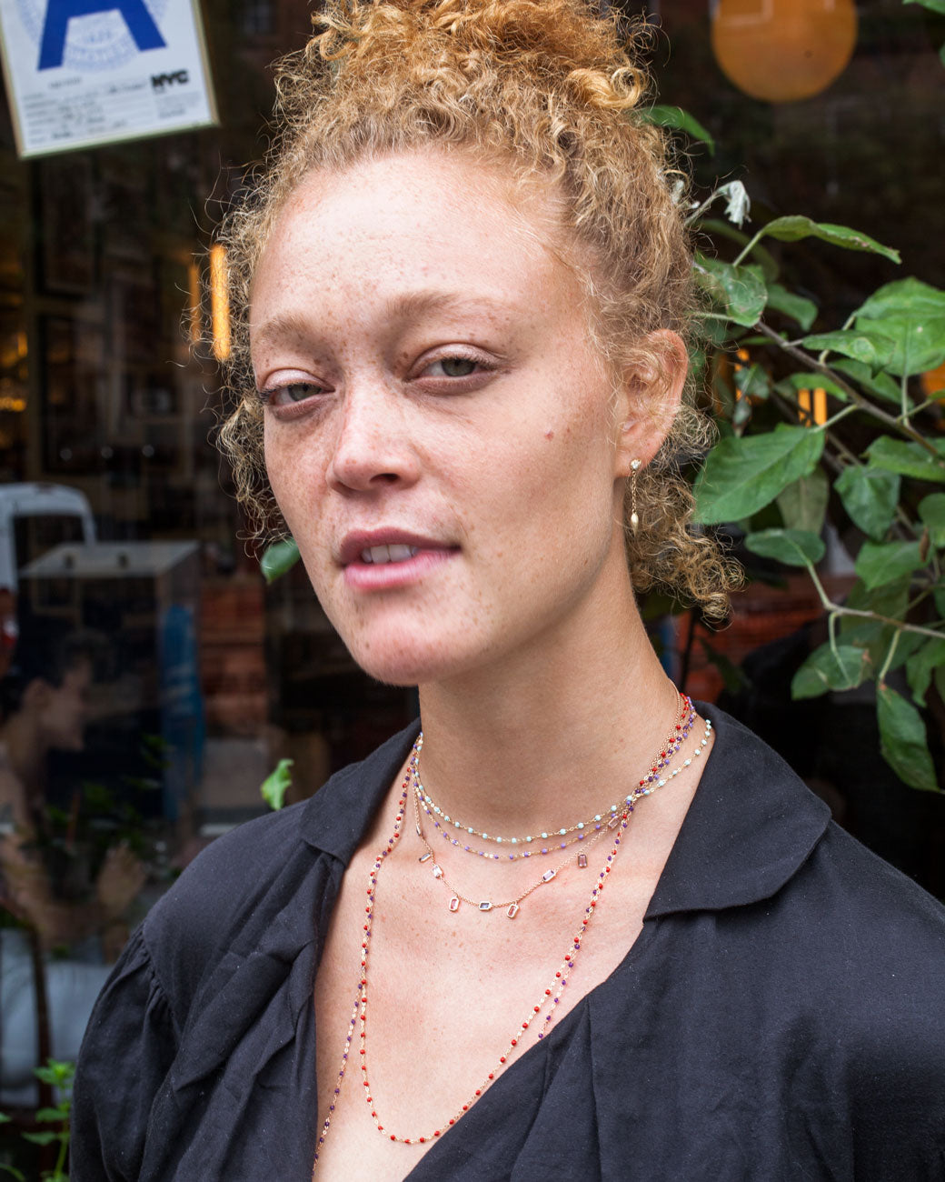 Image of woman with hair in a bun wearing black shirt and several yellow gold chain necklaces with resin beads of varying lengths, in addition to a yellow gold necklace with precious gemstones of varying colors.